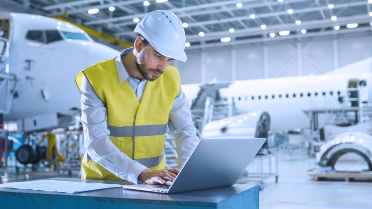 Ein Luftfahrtexperte mit Warnweste und Helm arbeitet in einem Hangar an seinem Laptop. Im Hintergrund sind Flugzeuge bei der Montage und Wartung zu erkennen.