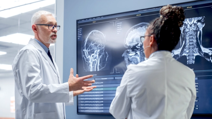 In einer Klinikumgebung bespricht ein Arzt mit einer Ärztin digitale MRT-Daten an einem großen Wanddisplay.