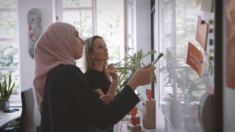 Eine Projektleiterin und eine Entwicklerin mit Kopftuch diskutierten eine IT-Architektur am Whiteboard.